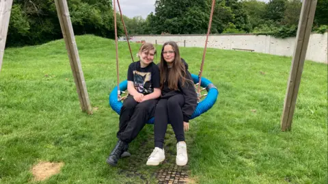 Kate Moser Andon/BBC Evie and Evie sitting next to each other on a big swing in the garden of Crofts Corner