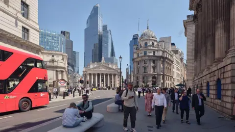 People walking through Bank junction in the sunshine and sitting on stone benches, a bus also travels through