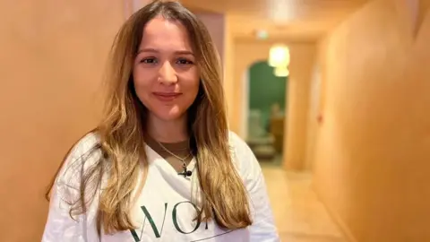 A woman with shoulder-length blonde hair looks at the camera smiling. She has a white t-shirt on.