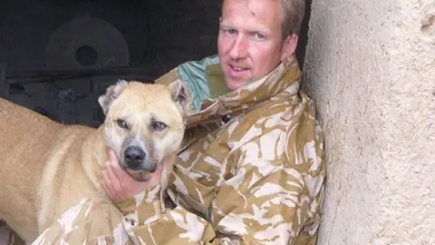 Nowzad Paul Farthing wearing light brown military fatigues while holding a dog with cream fur. Farthing is leaning against a wall.