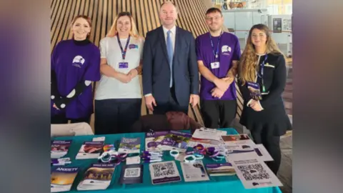 Papyrus Five people stand round a table covered in promotional leaflets, brochures, wristbands and pens. They are smiling to camera. Two of them are wearing purple t-shirts with the charity's logo on. 