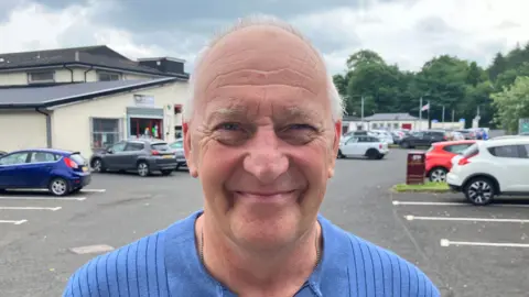 Mr Gurney is bald with grey hair and is wearing a blue top. The picture is taken in a car park with a number of cars parked. He is smiling at the camera.