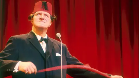 BBC Tommy Cooper, seen here in a fez performing one of his magic tricks. Behind him is a red curtain.