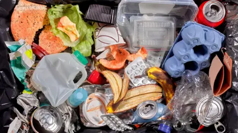 A bin filed with household waste, such as empty milk bottles and banana skins