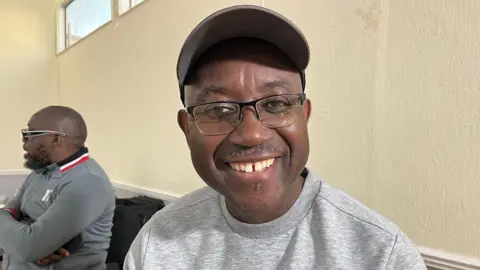 Nicola Haseler/BBC Dr Trevor Adams, in a room, wearing a grey jumper, glasses and a cap. Another man is sitting on a chair, to his left, with his arms crossed over his body. The walls are cream. Adams is smiling and looking at the camera. 