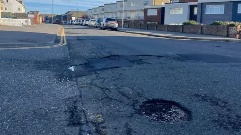 A pothole in the foreground and section of road behind which has a patched up bit of road in Seaford