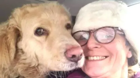 A middle-aged woman wearing glasses sits smiling with her face side-by-side with a dog's face