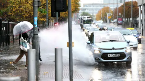 Pacemaker A man and woman are walking on a pavement in Belfast City Centre. It is raining and she is holding a clear umbrella over her head. The man has his hood up. A light blue car on the street is driving through a very large puddle and the water on the road is splashing up and hitting the couple on the pavement. 