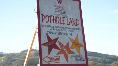 BBC A large hand-painted wooden sign with sunlit hills behind it. It reads Wrexham Council proudly presents Pothole Land, featuring two kilometers of award-winning potholes and very little actual road to spoil your fun. Guaranteed to be the deepest, longest and widest pothole in Wales. The darkest, longest and widest words read, spread across the orange and yellow star shapes. An arrow with the words, like this, points in the direction to the left. 