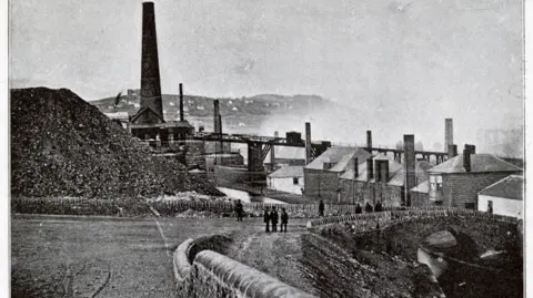 Alamy An historic image shows a range of historical industrial buildings at the copperworks site, with smoke seen in the background and about eight people seen standing near a little bridge over the river in the foreground.