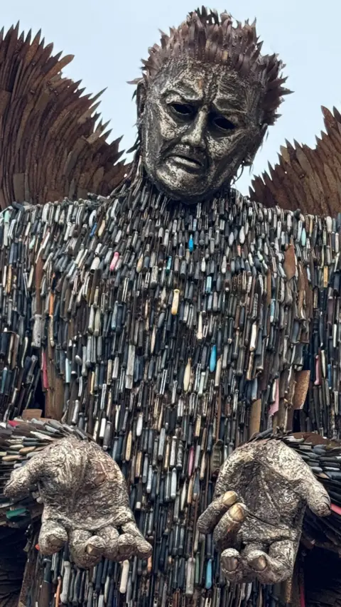 A statue of an angel with its hands out. It is made of knives and metal. There is a blue sky in the background. There is a BBC logo in the top left. 