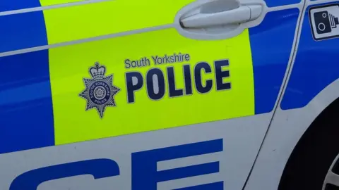 A close-up photograph of a South Yorkshire Police vehicle. The image focuses on the side door of a marked police car. In the centre is the text South Yorkshire POLICE alongside the force crest.