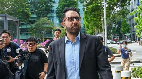 Pritam Singh wearing a suit and walking on a road. Photographers dressed in black are walking behind him.