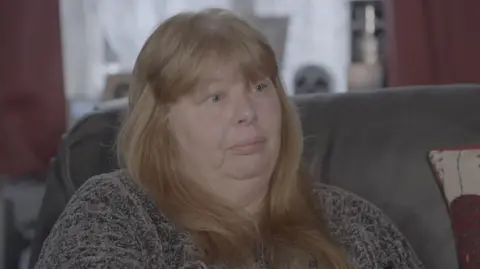 A woman with long and red blond hair sitting on a gray sofa, wearing a gray flying and looking forward to her left, because she is in a conversation with a BBC correspondent outside the camera