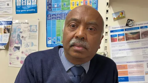 Nicola Haseler/BBC Bharat Shah sits in a room with medical posters in the background. He has a grey moustache and is wearing a blue checked shirt with navy tie and navy sweater on top. 