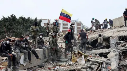 Rescuers search for victims at a collapsed building as one raises a Venezuelan flag following a magnitude 7.2 earthquake that struck Venezuela.