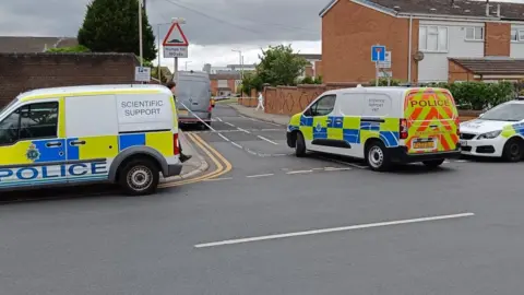 A police scientific support van is parked on the road and is on the left of the image. Two other police vehicles can be seen. Inside the cordon in the distance a forensic officer is searching the scene.