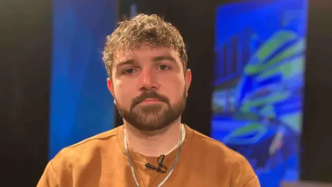 Fen Trudgian, wearing a tan jumper and a solid neck chain. He has a groomed beard and moustache and brown, lightly curly hair. He is looking at the camera and behind him there is an out of focus blue background.
