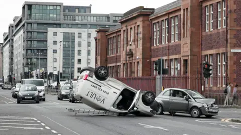 A white van with a ladder on top flipped on it roof with a small grey car with a smashed bonnet beside it.