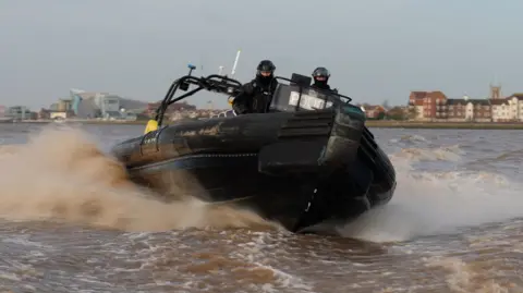 A black police rigid‑hulled inflatable boat moves at high speed, lifting slightly at the bow as it cuts through the water. Two officers in black protective gear and helmets are positioned near the front, bracing themselves as spray rises around the vessel. Behind them, on the far riverbank, rows of modern apartments and taller buildings stretch along the shoreline.