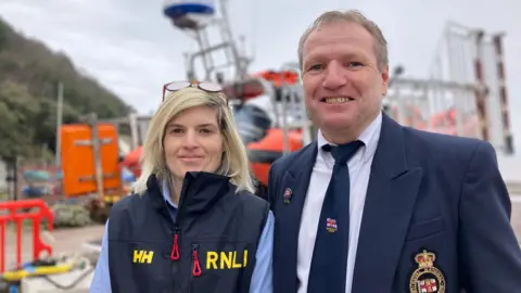 Karla is on the left, wearing an RNLI gilet and blue shirt, smiling at the camera. She has shoulder length blonde hair and glasse son her heard. On the right is Andrew in a blue RNLI blazer and tie, also smiling at the camera. Behind them are boats and trussing.