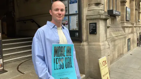 Jack Abraham holding a blue sign reading "More IRL, less URL" in front of Oxford Town Hall.