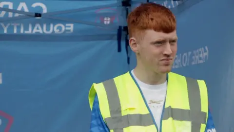 A man with red hair, wearing a hi-vis vest over the top of a Carlisle United football shirt.