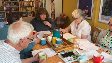 Ginny Sealey with members of a book club sat around a table. Ginny is at the head of the table in a blue jumper. There are two women and two men sat with her. There are pieces of paper and cups of tea on the table.