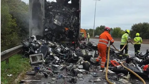 National Highways Firefighters battle the flames of a lorry fire on the M5 in Exeter. Two crew members are inspecting breathing apparatus on the road while others work on the fire.