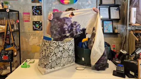A man holds up a purple and cream scarf with a hole in it, with his face partially visible through the hole. He is standing behind the counter of of a charity shop, with two bags of donations on the table in front of him. 