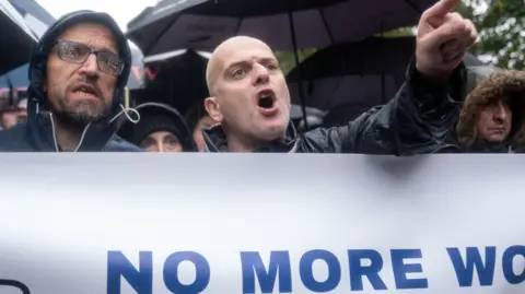 PA Media Image shows two men standing behind a banner saying 'NO MORE WORDS'. One is pointing a finger and shouting. 