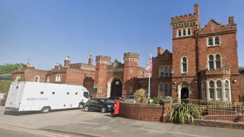 A large red‑brick prison building with castle‑like architecture, including towers and arched entrances. A white custody transport vehicle is parked in front, alongside several cars. A flag of Great Britain flies on a flagpole near the entrance, and a red post box is positioned by a low brick wall. The sky is blue.