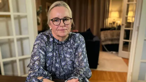 Amanda Ford sits at a table in her house. Her blonde hair is pulled back into a ponytail and she has black thick-rimmed glasses. She is wearing a blue and white paisley blouse,