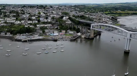 BBC Aerial view of two bridges spanning a river with boats moored in the river and houses on the bank of the river with fields beyond.