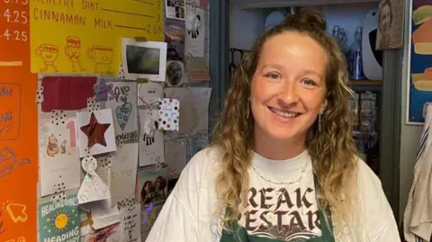  A woman is standing behind a counter in a colorful café-style setting with bright wall art, handwritten menus, and shelves holding jars of snacks in the background. She has long blonde hair and is smiling at the camera and is wearing a white T-shirt with bold writing on it and a green apron over it.

