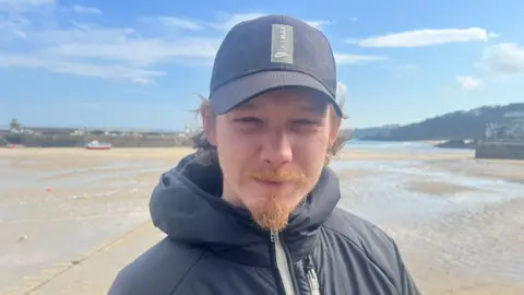 A man wearing a black cap and black coat, with a goatie beard, standing in front of the harbour
