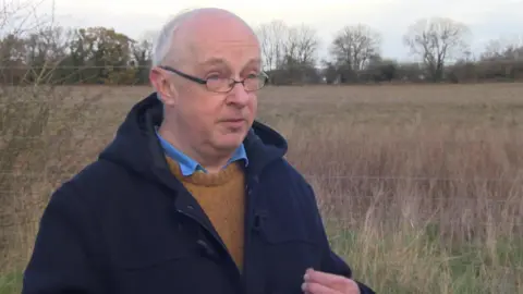 A man with short, grey hair and dark-rimmed glasses, wearing a brown jumper, blue shirt and a navy woollen coat.