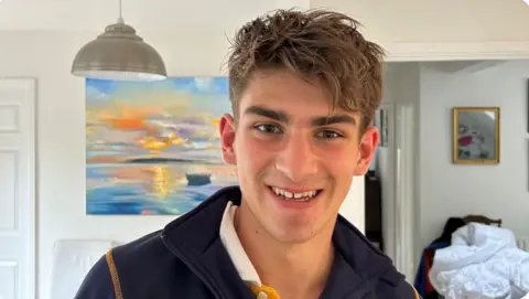 BBC Tom Dunlop is smiling at the camera. He has brown eyes and hair, is wearing a navy sweatshirt.