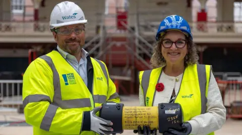 Derby City Council Tim Wates, from construction firm Wates and Derby City Council leader, Nadine Peatfield, holding a yellow time capsule inside Derby Market Hall