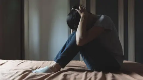 Anonymised generic photo of a woman sitting on a bed in her bedroom with her head in her hands. She is wearing a grey t-shirt and blue jeans and light coloured trainer socks and the bed sheet is light brown. 