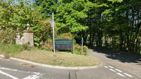 A black road sign for Inverness Crematorium and Kilvean Cemetery at the edge of a road - on a sunny day with green trees in the background.