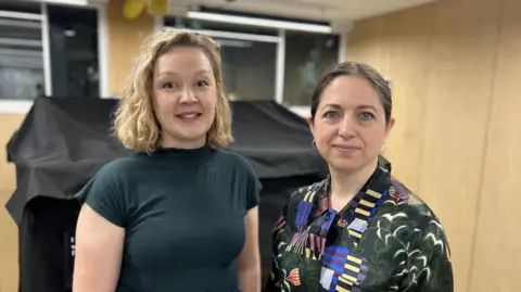 Woman on the left wears a black short sleeved top, has bobbed blonde hair and the woman next to her has a patterned dark blouse and dark brown hair tied back from her face. Behind them are light brown walls.