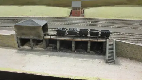 Bob Hetherington A building is in the foreground with five trailers on the tracks, two filled with coal. There are steps leading down from the track. 