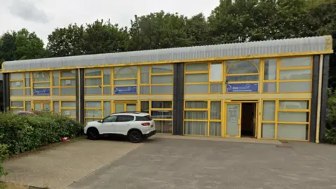 Google A Google Maps Street View image of the outside of the Ross Coates Solicitors building. Its frontage is yellow and predominantly features glass windows. It is a unit on an industrial estate. There is a white car parked outside.