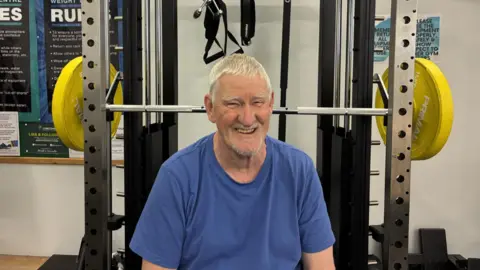 Gary Savage sits at a weight lifting bench with a barbell behind him. He is wearing a blue t-shirt and has white hair and a stubbly goatee beard.