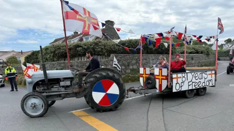 A tractor pulling a trailer. The tractor has red, white and blue wheels and a large Guernsey flag. The trailer has two people in it, two Guernsey flag shields and a sign which reads "81 years of freedom". 