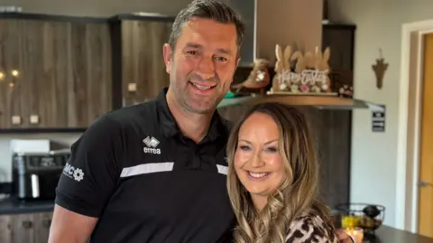 Graeme and Gemma Lee embracing in their kitchen. He is wearing a black polo shirt and she is wearing a leopard print top. Both are smiling and looking very happy.