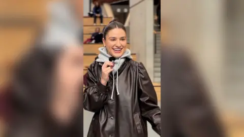 Student in a brown leather jacket with a grey hoody underneath. She has brown hair tied back and is holding a small microphone.