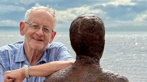 Ron Davies with grey hair wearing a blue and white shirt and glasses leans on one of the Iron Men sculptures on Crosby beach with the incoming tide in the background on a clear day. He is smiling. 
