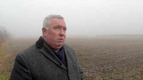 A man in a grey coat is standing on the edge of a muddy field. It is a foggy day but the outline of hedges can still be seen in the distance.